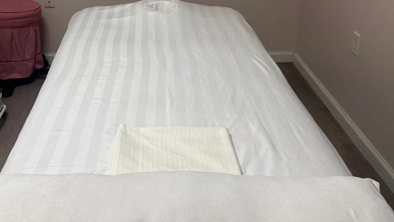 Massage table with white linens and a folded towel in a treatment room at Jin’s Massage in Glenside